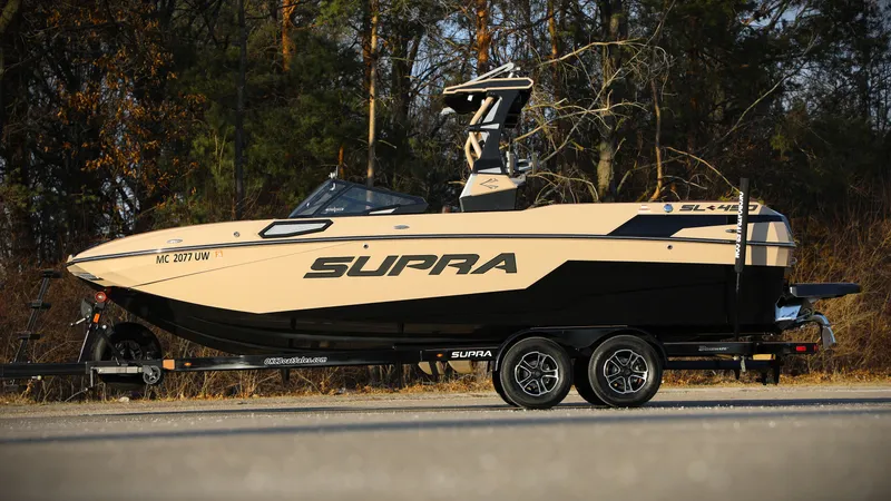 The Image of 2023 Supra SL boat on trailer, parked outdoors with forest background. - 0