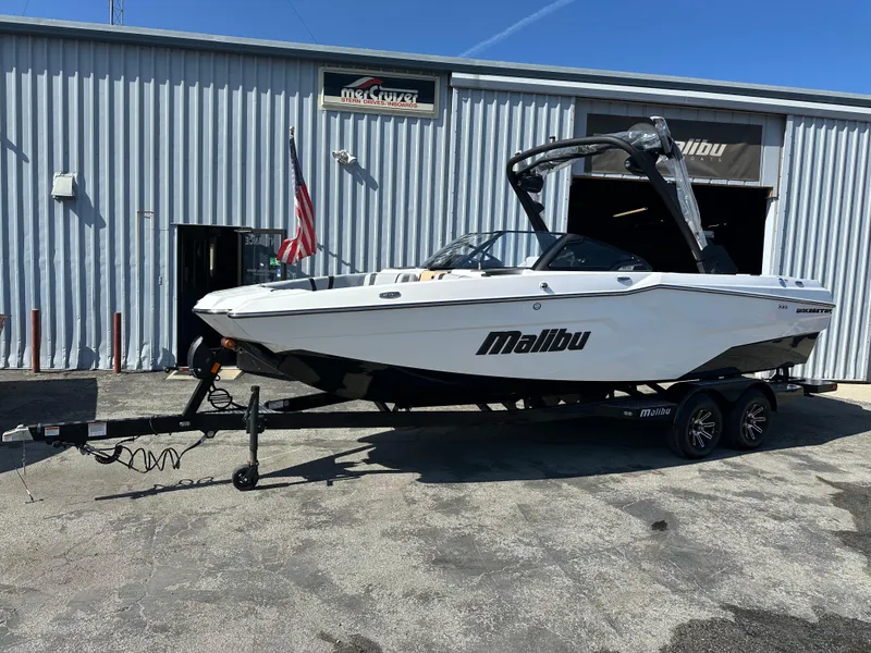 Slide: The Image of 2025 Malibu 23 MXZ boat on trailer outside a metal building, sunny day. - 1