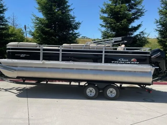 The Image of 2025 Sun Tracker SportFish 22 DLX pontoon boat on trailer, sunny day, surrounded by trees. - 0