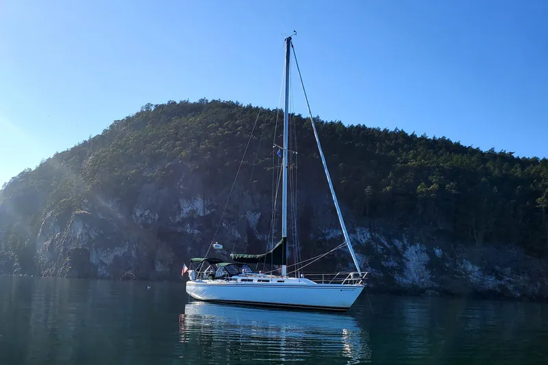 Slide: The Image of 1986 Catalina 36 sailboat anchored near a forested cliff under a clear blue sky. - 2