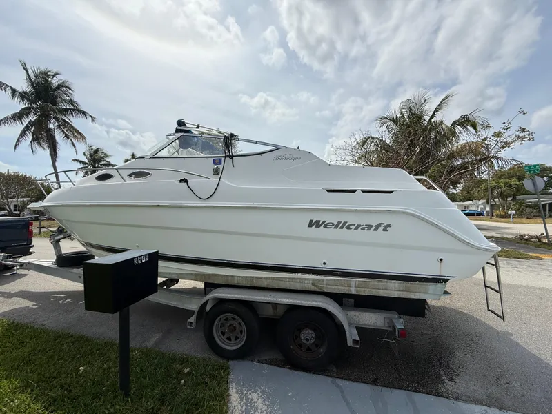 Slide: The Image of 2000 Wellcraft 2400 Martinique boat on trailer, parked near palm trees under cloudy sky. - 2