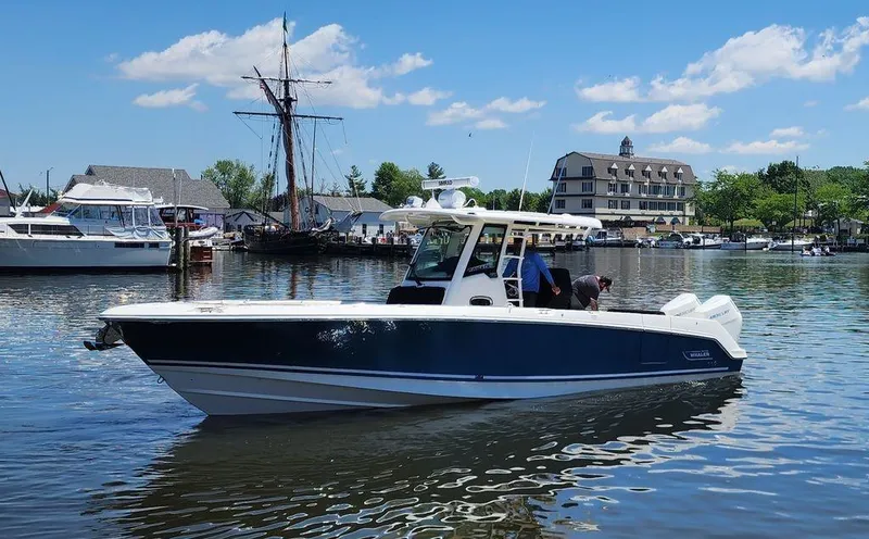 Slide: The Image of 2024 Boston Whaler 330 Outrage boat on calm water near a marina, under a clear blue sky. - 0