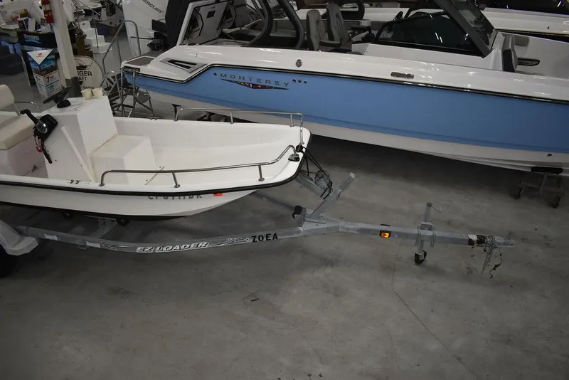 Slide: The Image of 2011 Carolina Skiff J16 boat on trailer, displayed indoors beside a larger blue and white boat. - 7