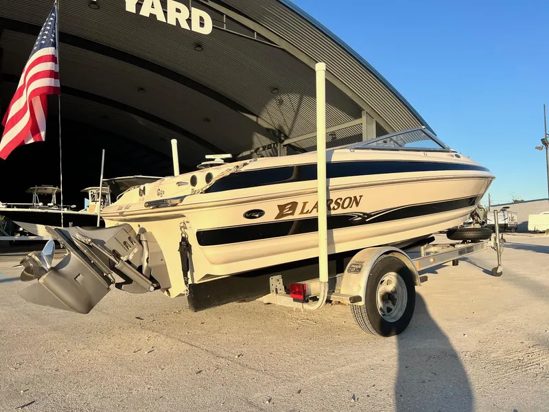 Slide: The Image of 2002 Larson LXi 190 boat on trailer, parked near a large building with an American flag. - 6
