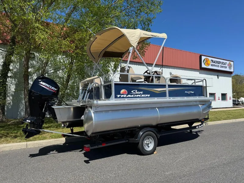Slide: The Image of 2025 Sun Tracker Bass Buggy 16 XL Select pontoon boat on trailer, parked near service center. - 2