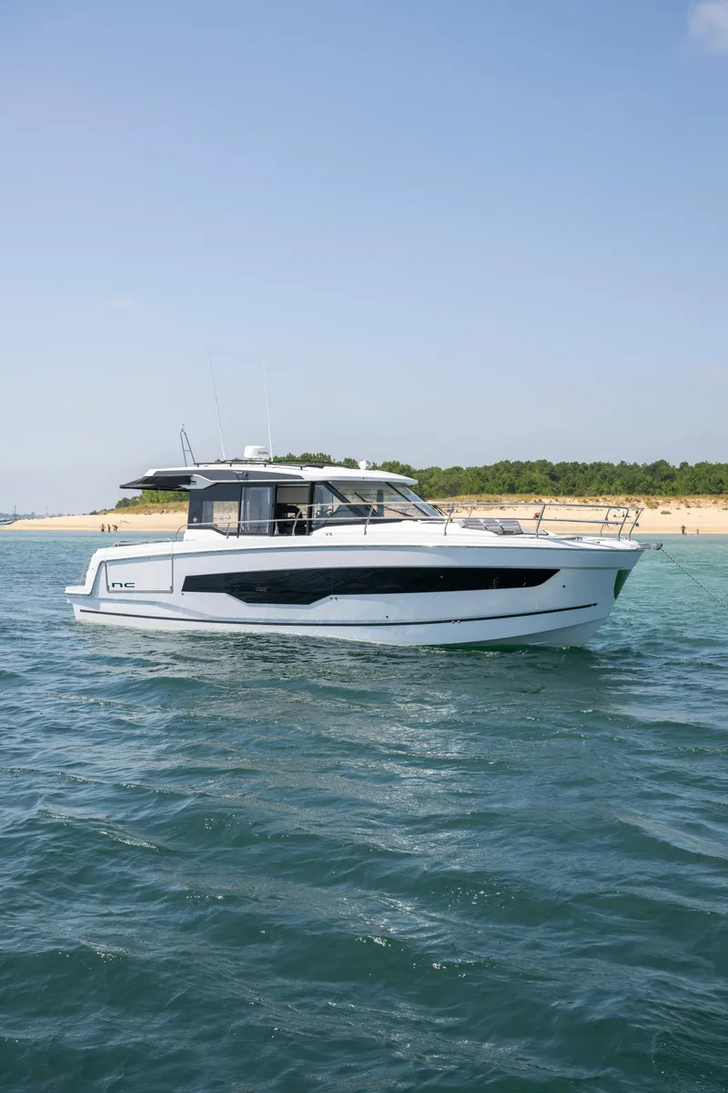 Slide: The Image of 2025 Jeanneau NC 1295 Coupe yacht cruising on open water near a sandy shoreline. - 6