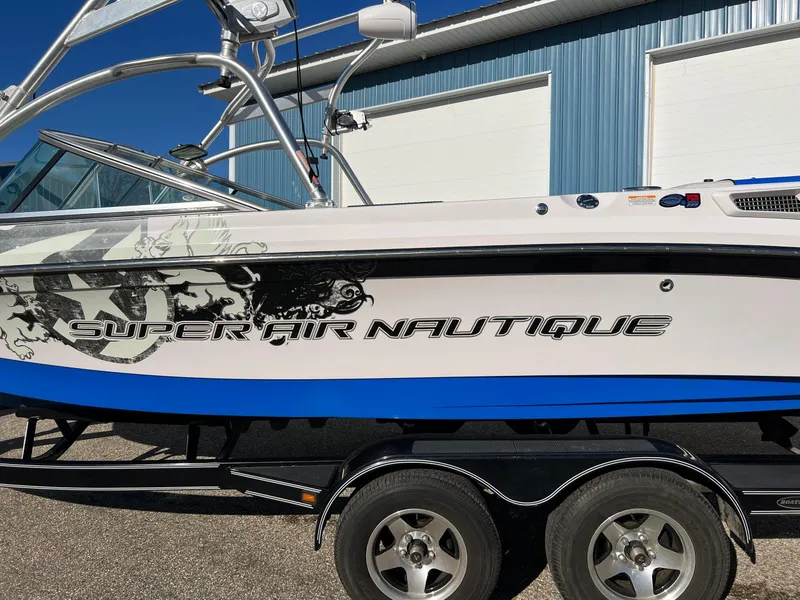 Slide: The Image of 2008 Nautique 210 boat with custom graphics, parked on a trailer near a blue building. - 13