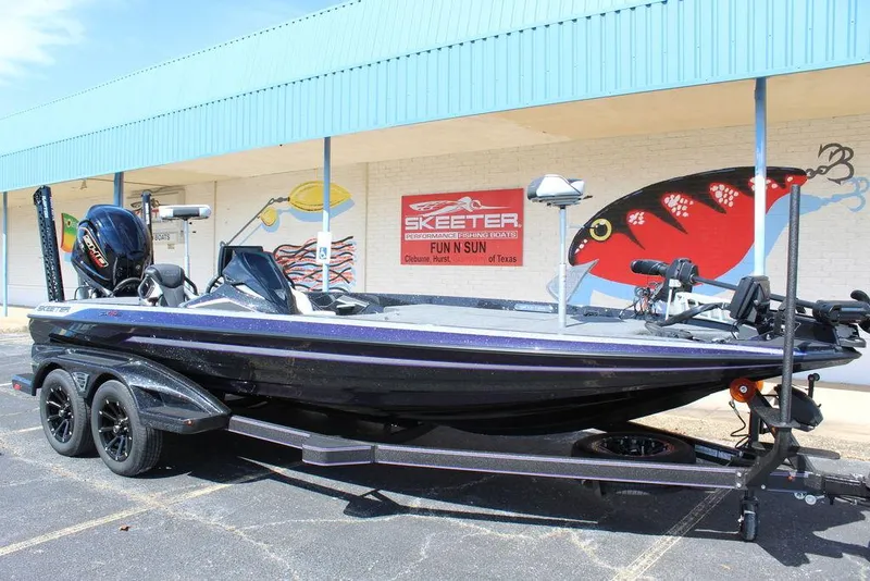 Slide: The Image of 2025 Skeeter FXR21L boat on trailer, parked outside a building with fishing-themed murals. - 1