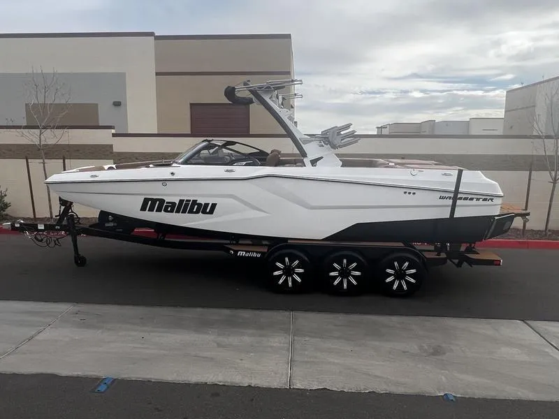 Slide: The Image of 2025 Malibu 24 MXZ boat on trailer, parked outdoors near modern buildings. - 1