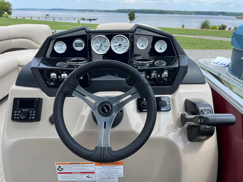 Slide: The Image of 2024 Landau Island Breeze 232 Cruise dashboard with steering wheel and controls, overlooking a scenic lake. - 13