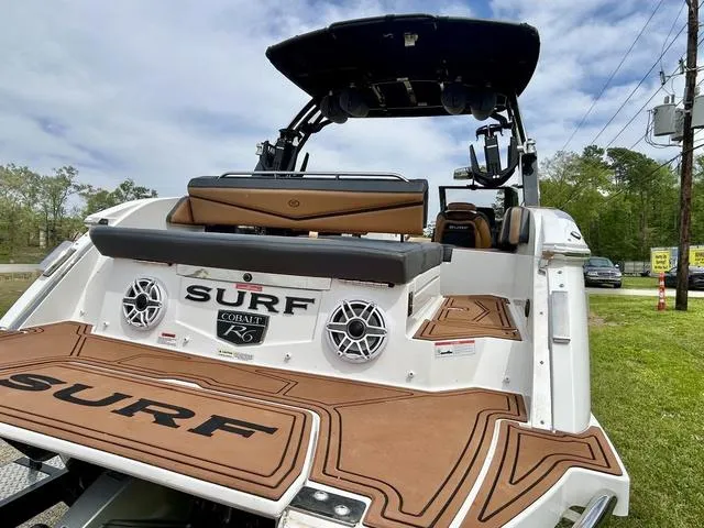 Slide: The Image of 2023 Cobalt R6 Surf boat with brown deck, speakers, and seating, parked outdoors. - 11