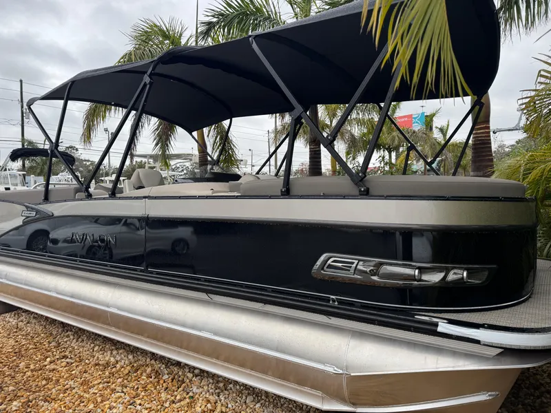 Slide: The Image of 2025 Avalon Catalina Quad Lounge pontoon boat with canopy, parked on gravel, surrounded by palm trees. - 6