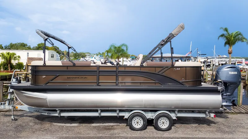 Slide: The Image of 2025 Bennington 22 SSR pontoon boat on trailer, dockside with palm trees and marina background. - 1
