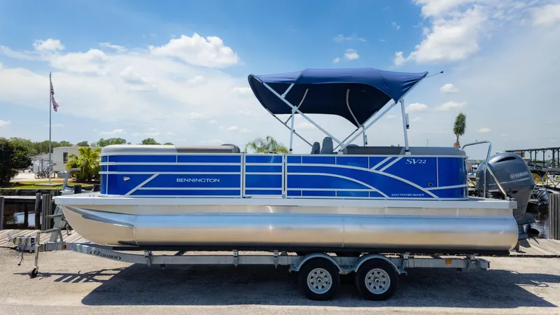 Slide: The Image of 2024 Bennington 22 SVSR pontoon boat with blue canopy on trailer, under clear sky. - 26