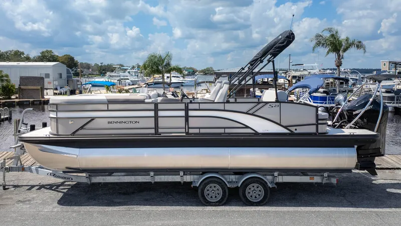 Slide: The Image of 2025 Bennington 23 SSB pontoon boat on trailer at marina, under a partly cloudy sky. - 1