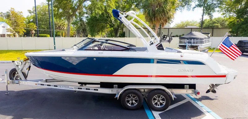 Slide: The Image of 2024 Cobalt CS23 Surf boat on trailer with American flag, parked outdoors. - 5
