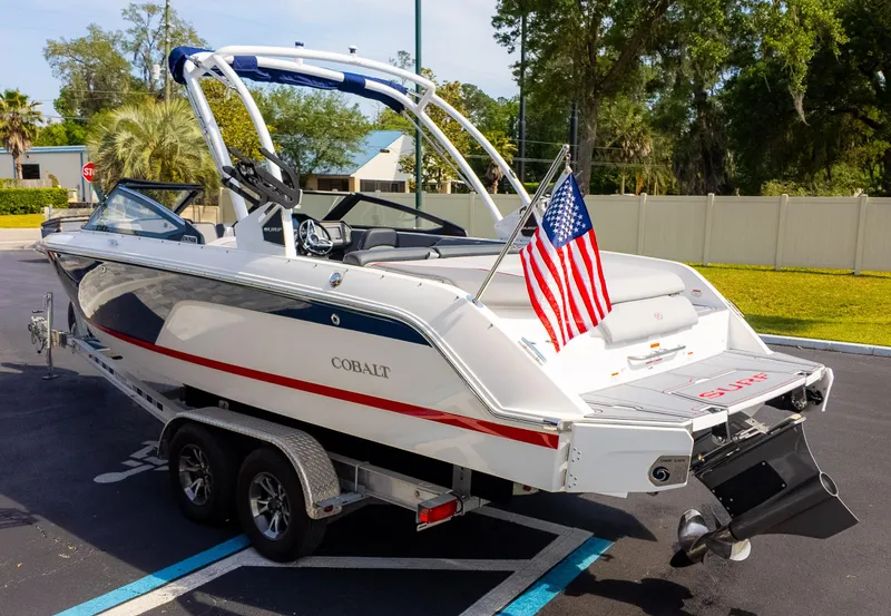 Slide: The Image of 2024 Cobalt CS23 Surf boat on trailer with American flag, parked outdoors. - 6