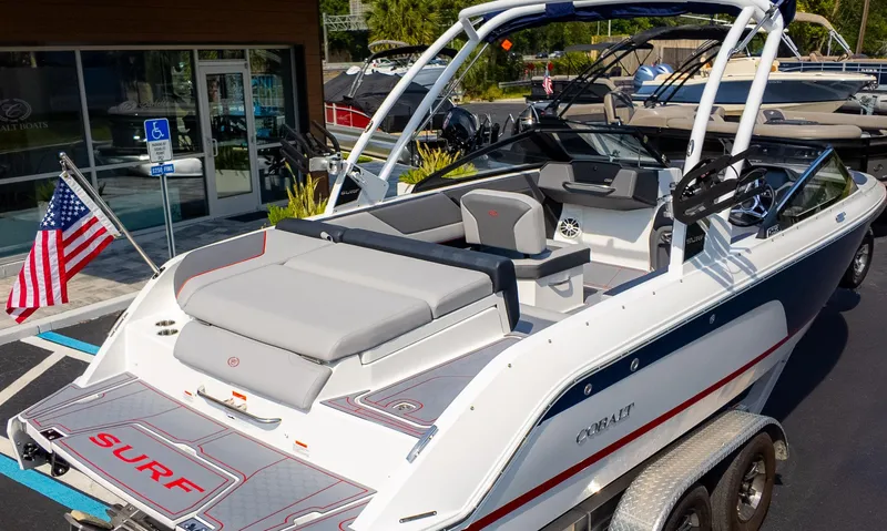 Slide: The Image of 2024 Cobalt CS23 Surf boat with American flag, parked on a trailer. - 8
