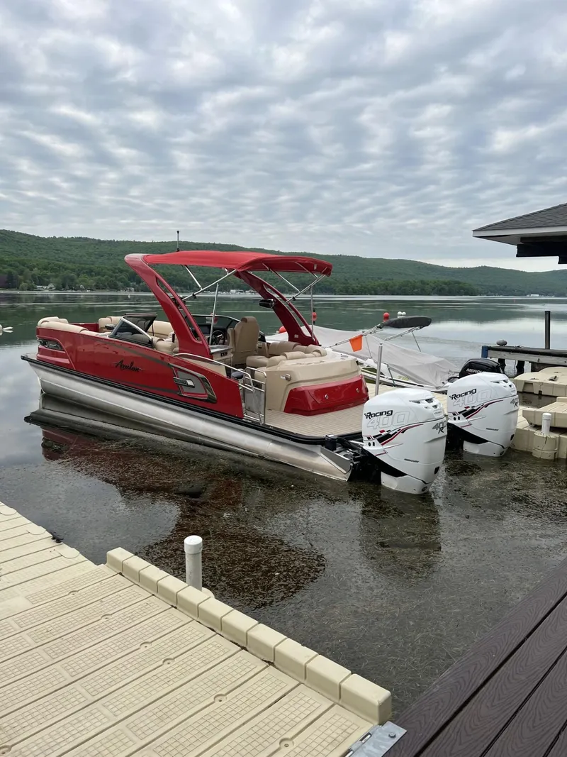 Slide: The Image of 2022 Avalon Excalibur Elite Windshield boat docked on a serene lake under a cloudy sky. - 2