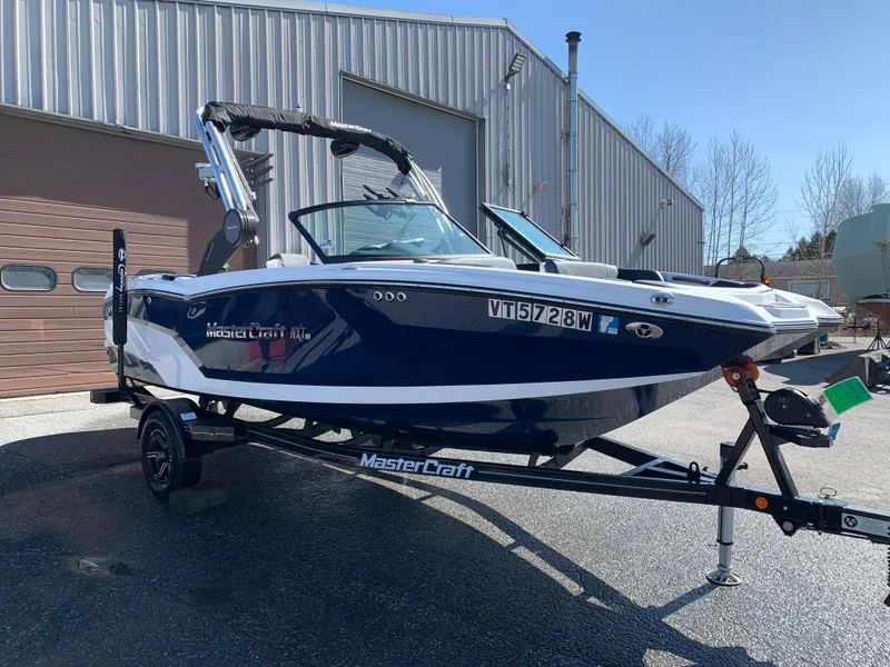 Slide: The Image of 2023 MasterCraft NXT20 boat on trailer, parked outside industrial building. - 0