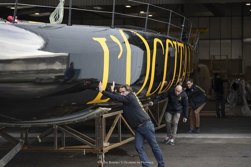 Slide: The Image of Men pushing Black Pepper Imoca 2025 yacht in a workshop. - 7