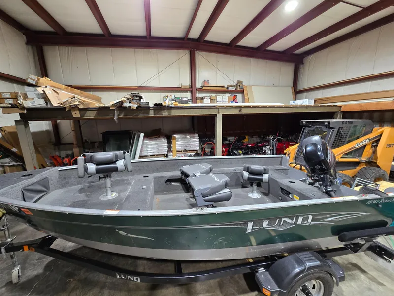 Slide: The Image of 2014 Lund 1650 Rebel Tiller boat in a warehouse, featuring a green exterior and trailer. - 2