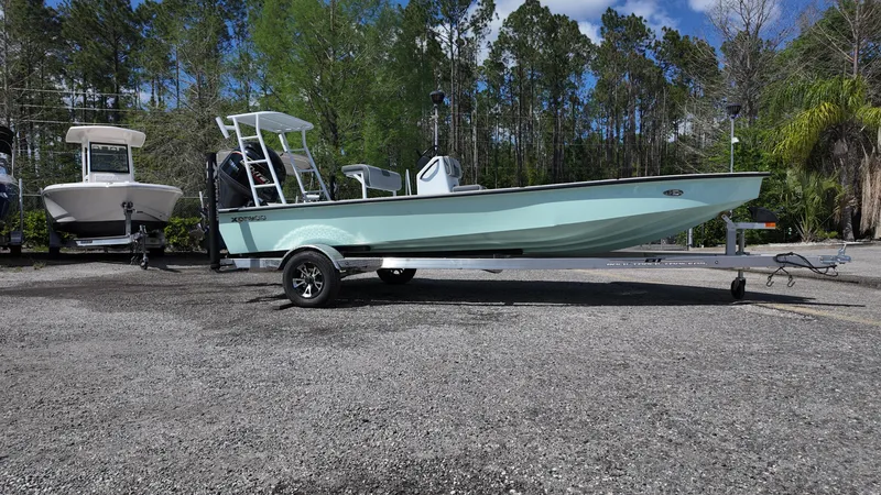 Slide: The Image of 2025 Xpress Skiff 185 boat on trailer, parked outdoors with trees in background. - 6