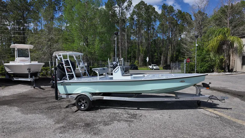Slide: The Image of 2025 Xpress Skiff 185 boat on trailer, parked outdoors near trees and other boats. - 7