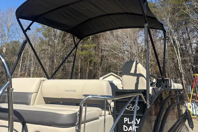 Slide: The Image of 2023 Harris HCX230 SL pontoon boat with canopy, beige seating, and wooded background. - 7