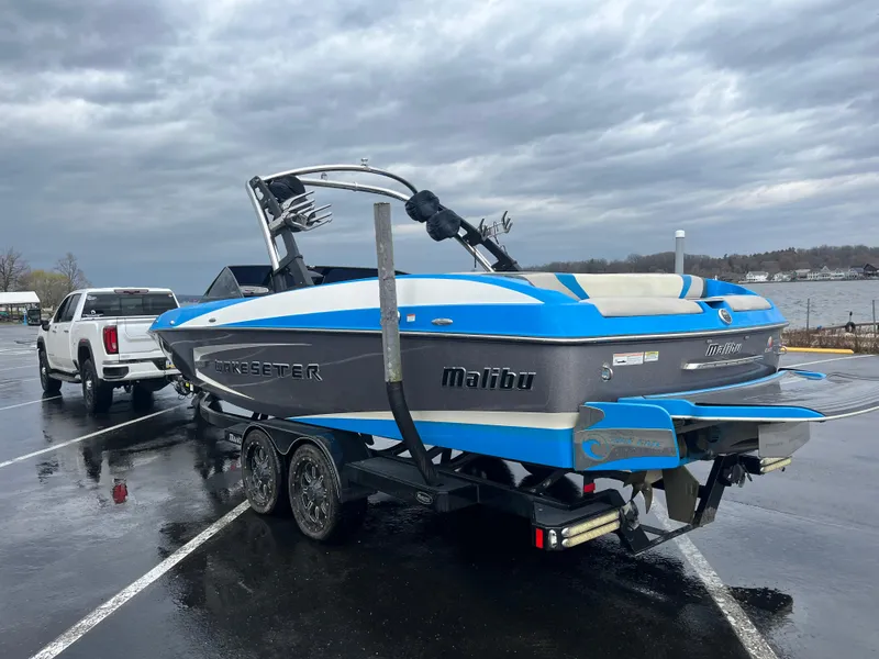 Slide: The Image of 2014 Malibu Wakesetter 24 MXZ boat on trailer, parked near a lake under cloudy skies. - 10