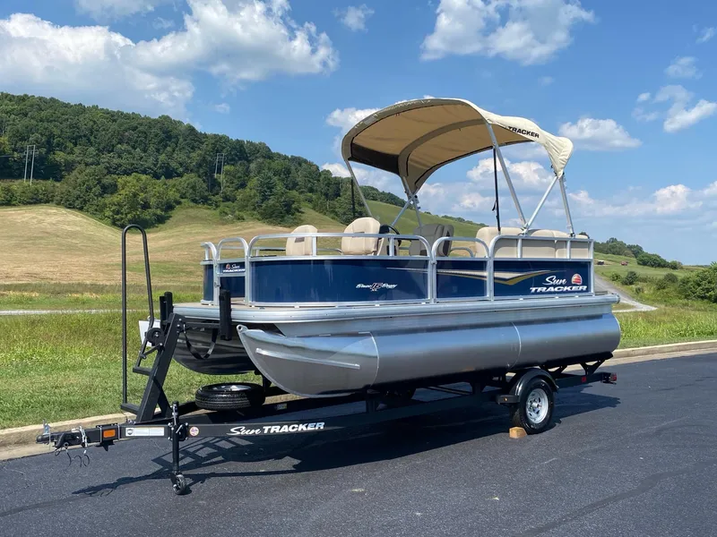 Slide: The Image of 2025 Sun Tracker Bass Buggy 16 XL Select pontoon boat on trailer, scenic background. - 5