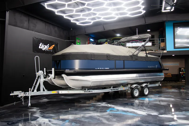 Slide: The Image of 2025 Viaggio Lago X-Q pontoon boat on trailer in showroom with modern lighting. - 1
