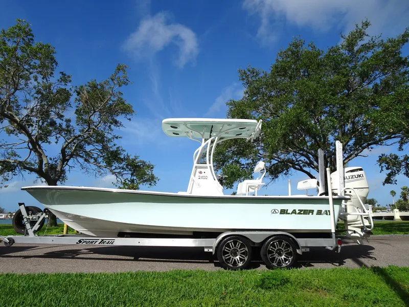 Slide: The Image of 2019 Blazer Bay 2400 boat on trailer, parked near trees under a clear blue sky. - 6