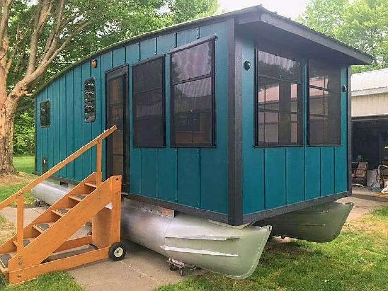 Slide: The Image of Custom 2023 houseboat with teal exterior, wooden stairs, and pontoon base in a grassy area. - 2