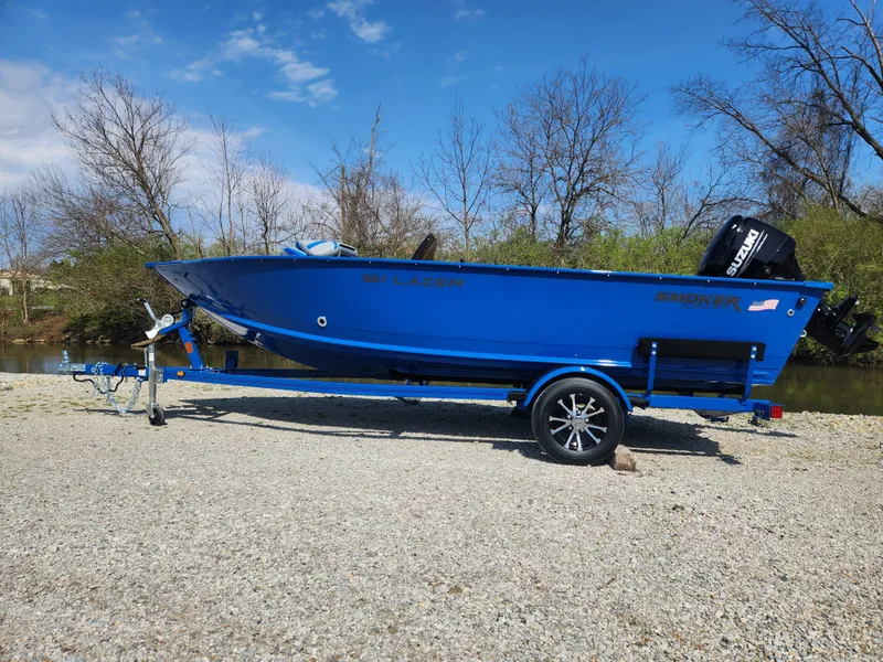 Slide: The Image of 2025 Smoker Craft Lazer 161 boat on trailer, blue exterior, parked on gravel near water. - 19
