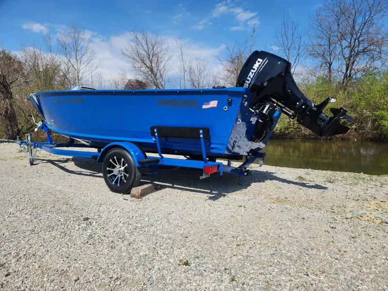 Slide: The Image of 2025 Smoker Craft Lazer 161 boat with Suzuki motor on a trailer by a river. - 2