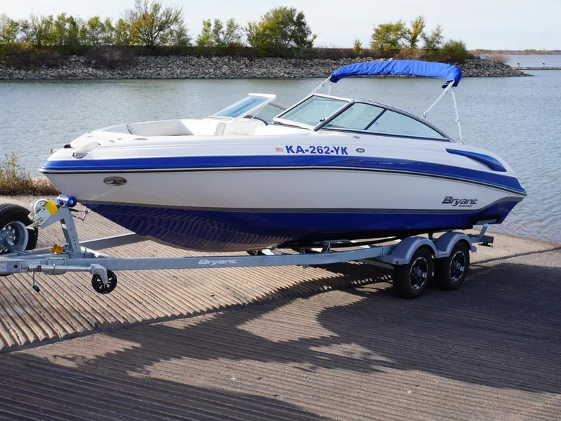 Slide: The Image of 2013 Bryant 220 boat on trailer by a lake, featuring blue and white design. - 0