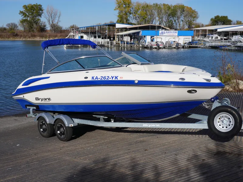 Slide: The Image of 2013 Bryant 220 boat on trailer by a lake, featuring blue and white design. - 1
