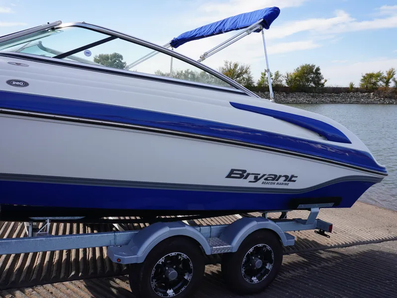 Slide: The Image of 2013 Bryant 220 boat on trailer by the water, featuring blue and white design. - 4