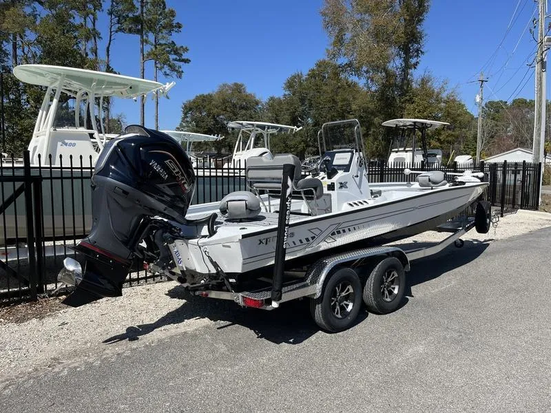Slide: The Image of 2025 Xpress H22B boat on trailer, parked outdoors, with trees and other boats in background. - 2
