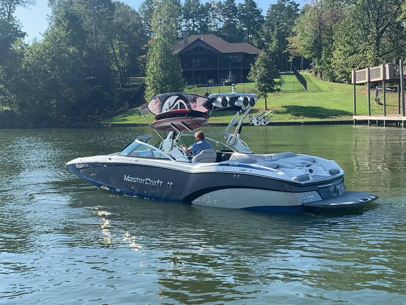 Slide: The Image of 2015 MasterCraft X23 boat cruising on a serene lake near a wooded shoreline. - 2