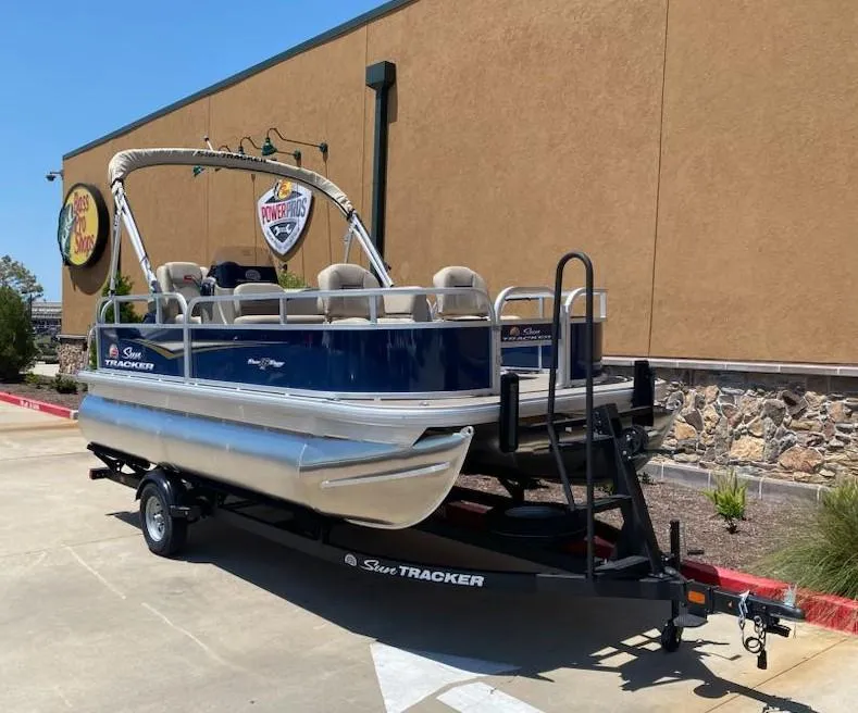 Slide: The Image of 2025 Sun Tracker Bass Buggy 16 XL Select pontoon boat on trailer, parked outside a store. - 2
