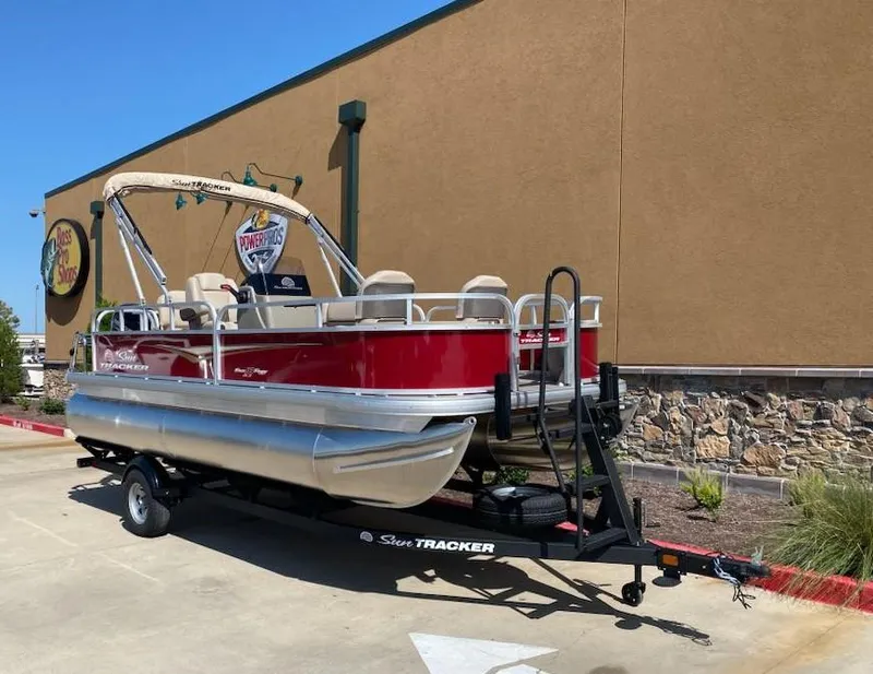 Slide: The Image of 2025 Sun Tracker Bass Buggy 18 DLX pontoon boat on trailer, parked outdoors. - 2