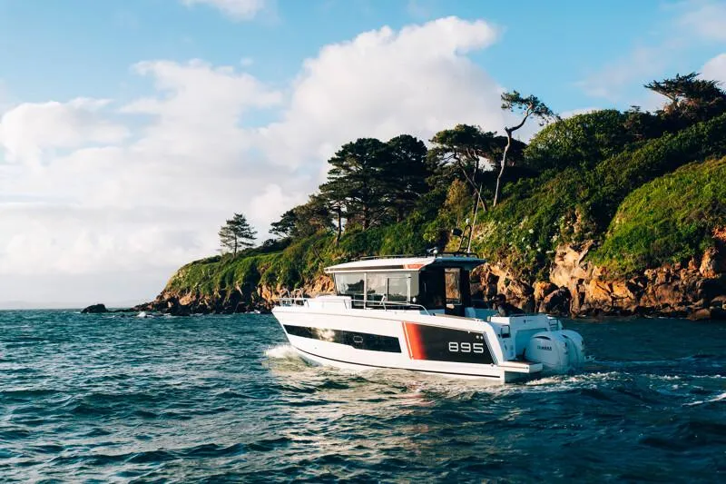 Slide: The Image of 2025 Jeanneau NC895 Sport S2 boat cruising near a scenic, tree-lined coastline. - 0