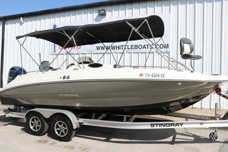 Slide: The Image of 2018 Stingray 212 SC boat on trailer with canopy, parked outside a building. - 12