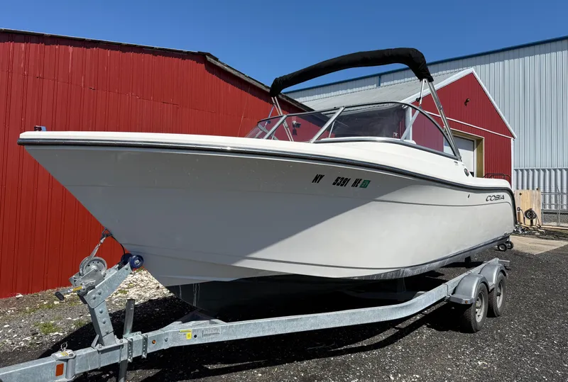Slide: The Image of 2023 Cobia 220 Dual Console boat on trailer, parked near red and gray buildings. - 1