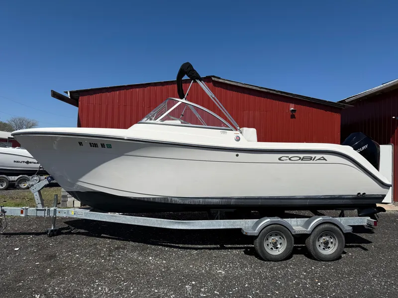 Slide: The Image of 2023 Cobia 220 Dual Console boat on trailer, parked near red building. - 2