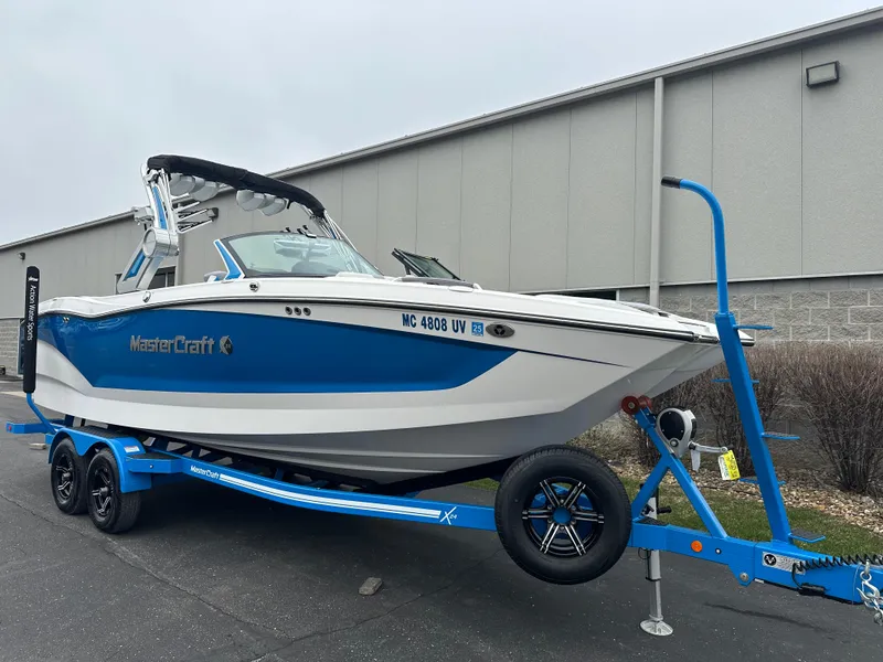 Slide: The Image of 2023 MasterCraft X24 boat on trailer, blue and white design, parked outdoors. - 2