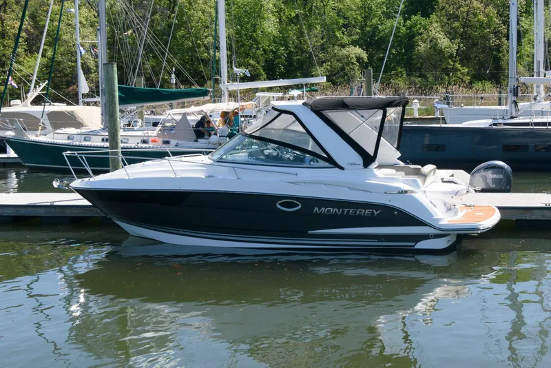 The Image of 2019 Monterey 295 Sport Yacht docked at marina, surrounded by sailboats and lush greenery. - 0