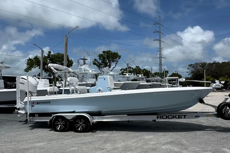 Slide: The Image of 2024 Contender 26 Bay boat on a Rocket trailer, parked outdoors under a cloudy sky. - 2